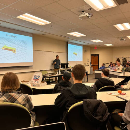 A university classroom features a lecturer standing at the front, speaking to a group of attentive students seated at curved rows of desks. Two large projection screens display a slide titled "First Steps" with a yellow 3D object on a grid.